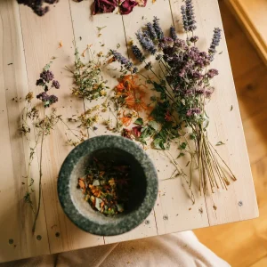 A person seated at a wooden table surrounded by various dried herbs, engaged in an activity related to herbal preparation