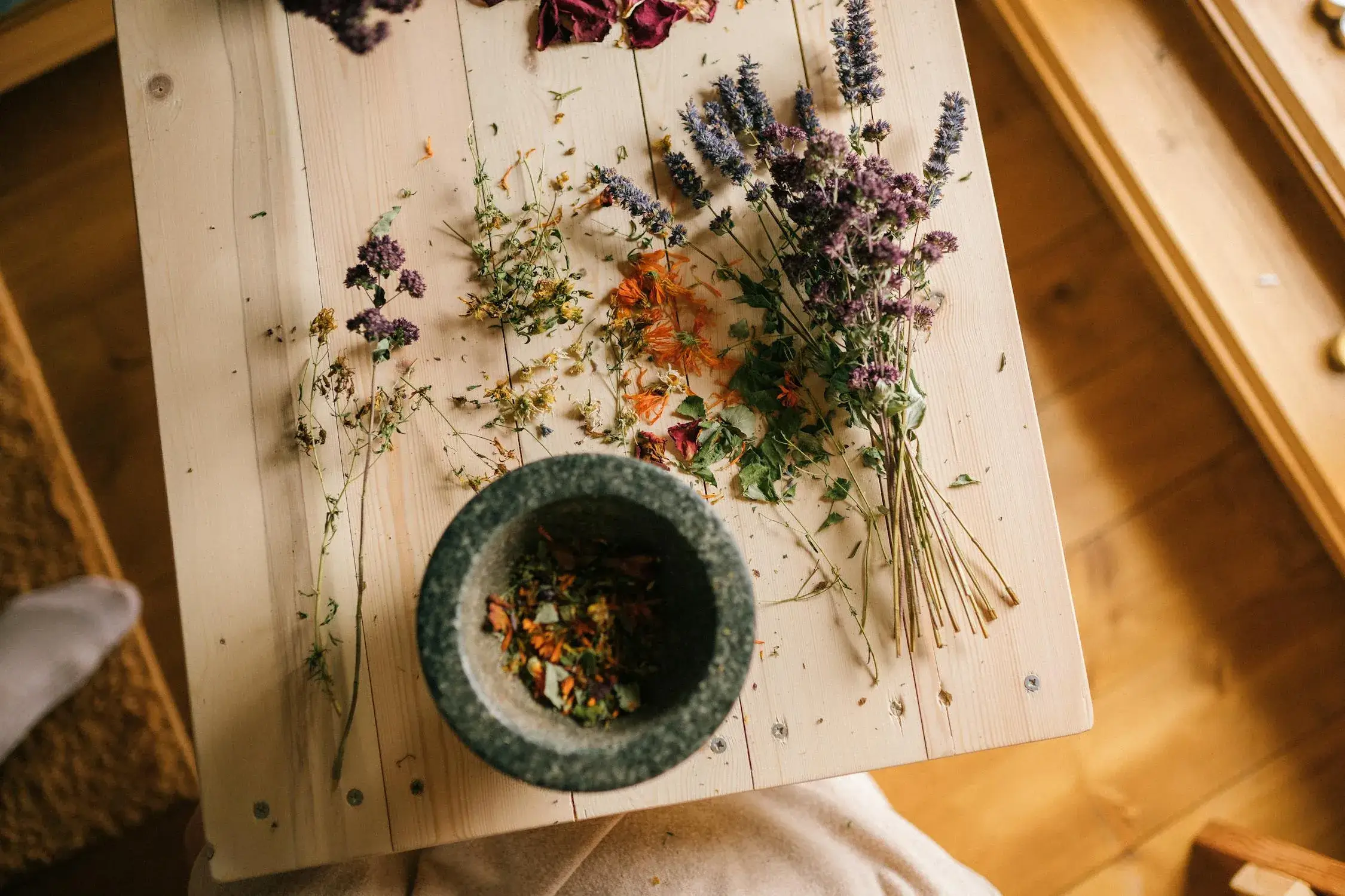mind and body 0 A person seated at a wooden table surrounded by various dried herbs, engaged in an activity related to herbal preparation